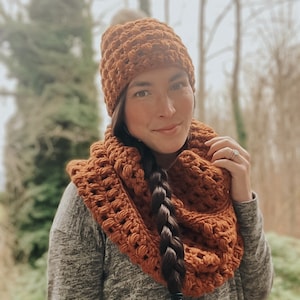 May include: A brown crocheted hat and matching infinity scarf. The hat has a pompom on top. The scarf is worn around the neck and drapes down the front of the wearer.