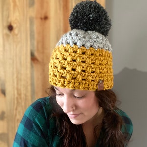 May include: A crocheted beanie with a yellow, gray, and black color scheme. The beanie has a pom-pom on top and a brown leather tag.
