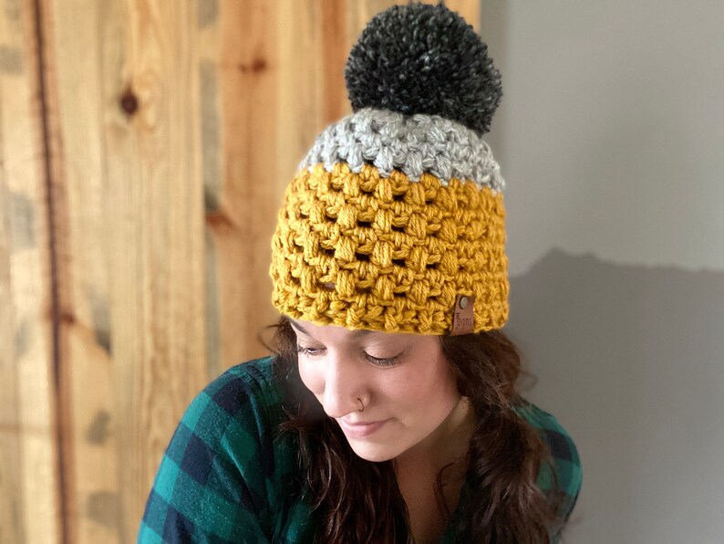 May include: A crocheted beanie with a yellow, gray, and black color scheme. The beanie has a pom-pom on top and a brown leather tag.