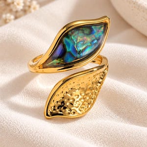 May include: A gold-toned open ring featuring two leaf-shaped ends. One end is inlaid with a polished, iridescent abalone shell displaying shades of blue and green, while the other end has a textured, hammered gold-toned surface finish.