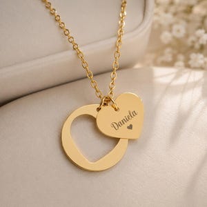 Puede incluir: Un collar de tono dorado con una cadena fina y dos colgantes: un aro circular y un dije sólido en forma de corazón. El dije de corazón tiene grabado el nombre Daniela y un pequeño símbolo de corazón, suspendido de la cadena.
