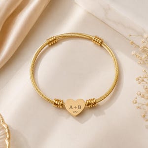 Puede incluir: Una pulsera rígida de cable dorado con un dije central en forma de corazón grabado con las letras A+B y un símbolo de infinito. La pulsera está decorada con cuatro cuentas cilíndricas estriadas a lo largo de la banda circular.