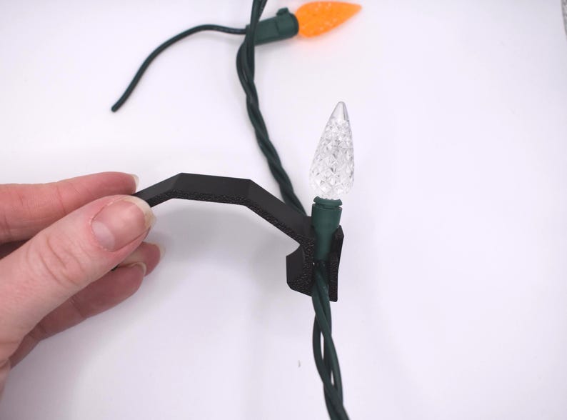 May include: A black plastic clip for securing Christmas lights to a surface. The clip is attached to a strand of green Christmas lights with white and orange bulbs.