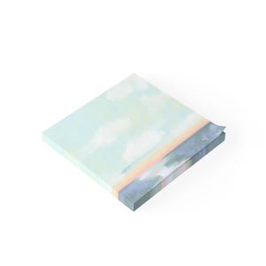 May include: A square notepad with a painted landscape design. The artwork features a sky with clouds, a colorful horizon, and a dark blue area at the bottom. The notepad is light blue and white.