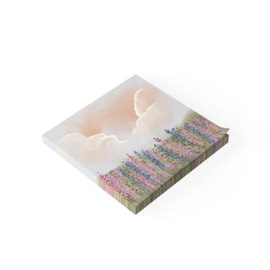 Puede incluir: Un bloc de notas cuadrado con una pintura de acuarela de un campo de lavanda bajo un cielo nublado. La paleta de colores incluye rosas, morados, azules y verdes suaves. El bloc de notas tiene una pila de páginas y los bordes son visibles.
