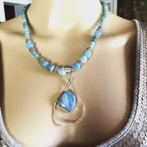 Peut inclure: Un collier de perles bleues avec un pendentif argenté. Le collier présente différentes nuances de perles bleues et un pendentif en forme de goutte d'eau avec une pierre bleu clair. Le pendentif est suspendu à un cadre argenté.