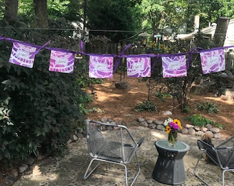May include: A purple and white banner with the words "peace" and "love" displayed on individual square flags. The flags are strung together with purple ribbon and hang in an outdoor setting. The background includes trees, a patio, and a small table with flowers.