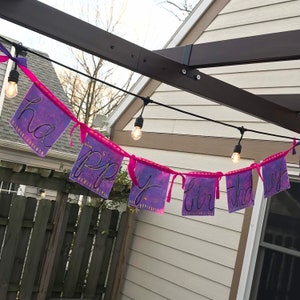 May include: A pink and purple banner with the words "Happy Birthday" written in black cursive. The banner is strung up with white string lights.
