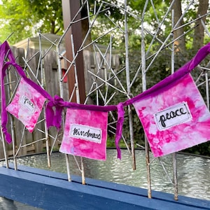 以下が含まれることがあります： 黒のマーカーで「grace」、「kindness」、「peace」と書かれたピンクとパープルの布の横断幕です。