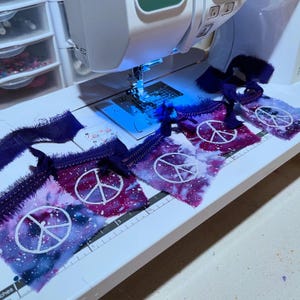 May include: A sewing machine with a purple fabric peace sign banner being sewn. The fabric is a tie-dye pattern with white peace signs.