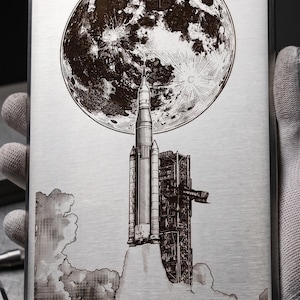 May include: A rectangular metal plaque featuring an engraved image of a rocket launching towards a large moon. The rocket is on a launchpad, with smoke and clouds. The moon is detailed with craters. Text at the bottom reads "SERVO-TECH ENGRAVING - SN: LT-001".
