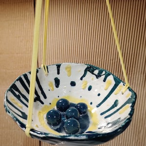 May include: A handmade ceramic hanging planter with a white interior, decorated with blue and yellow accents. The planter has a unique, irregular shape and contains several dark blue glass marbles. Yellow cords suspend the planter.
