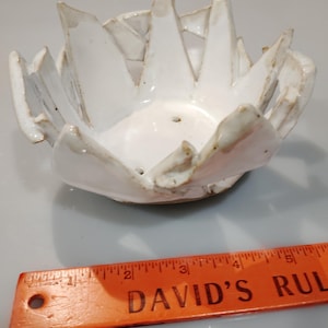 May include: A handmade white ceramic bowl with a unique, textured design. The bowl has a series of outward-facing, pointed edges, creating a starburst effect. The bowl is approximately 6 inches in diameter.