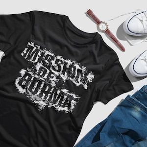 May include: Black t-shirt with a white distressed graphic that says "Mission de Burma". The shirt is laid out on a white surface with a pair of white sneakers, blue jeans, and a brown watch.