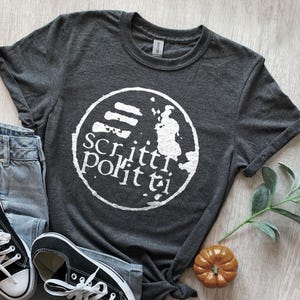 May include: A dark gray t-shirt with a white circle graphic that says "Scriitti Por it ti" and a stylized image of a person holding a paintbrush.