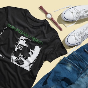 Op de afbeelding: Zwart T-shirt met een witte afbeelding van een gezicht met de tekst "SUBURBAN LAWNS" in groen.