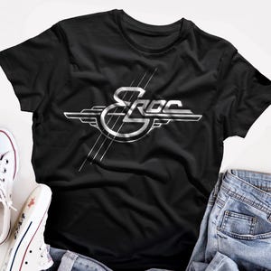 Peut inclure: T-shirt noir avec un logo argenté "SROC" au design stylisé. Le motif comprend des lignes et des formes, lui donnant une esthétique rétro. Le t-shirt est associé à des baskets blanches et un jean bleu. Le tissu est doux.