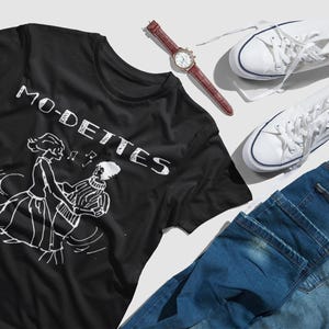 Pode incluir: Camiseta preta com um gráfico branco de duas pessoas dançando e o texto "MO-DETTES" impresso na frente.