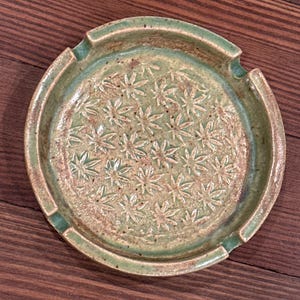 May include: A green ceramic ashtray with a floral design. The ashtray has a raised rim and a small indentation for cigarettes.