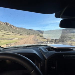 May include: Interior view from a vehicle, showing a dirt track through a desert environment. The steering wheel and dashboard are in focus, with a tablet device on the dash. Mountains and a clear blue sky are visible in the distance.