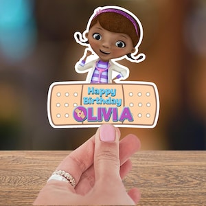 May include: A colorful birthday decoration featuring a cartoon character and the text "Happy Birthday OLIVIA". The design includes a cartoon image of a girl in a doctor's coat and a bandage shape with the text.