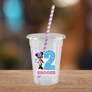 May include: A clear plastic tumbler with a lid and a purple and white striped straw. The cup features a graphic of a cartoon mouse wearing a pink bow and heart-shaped sunglasses, a large blue number two, and the name BROOKE in pink letters.