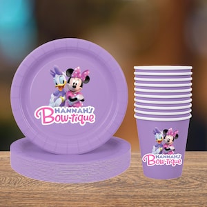 May include: A stack of purple paper plates and a stack of purple cups. The plates and cups have a cartoon image of Minnie Mouse and Daisy Duck. The text "Hannah's Bow-tique" is printed on the plates and cups.