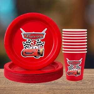 May include: Red party supplies featuring Lightning McQueen from Cars. Includes plates, cups, and a stack of plates. The plates and cups have the number 3 and the name Trevor.