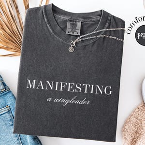 Op de afbeelding: Donkergrijs t-shirt met de witte woorden "MANIFESTING a wingleader". Een zilveren ketting met een cirkelvormige hanger ligt op het shirt. Het shirt ligt naast een spijkerbroek en witte sneakers.