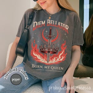 May include: A charcoal gray short-sleeve t-shirt featuring a graphic print of a sword piercing a crown, surrounded by red flames and a crescent moon. The text reads DIEM BELLATOR, BURN, MY QUEEN, and GLOW SO BRIGHT, THE DARKNESS TREMBLES.