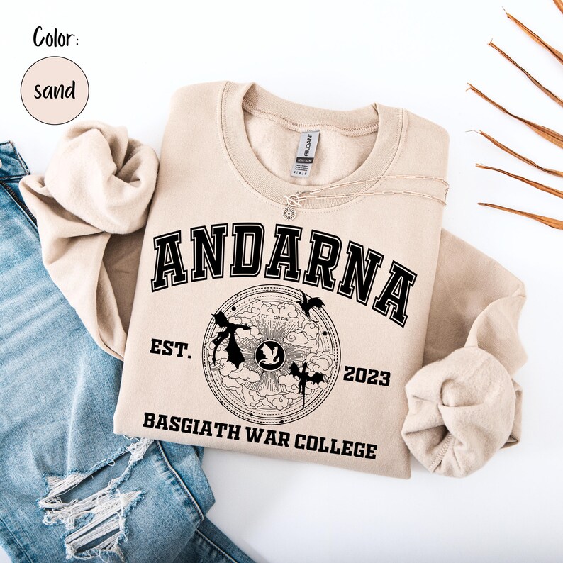 May include: Sand-colored sweatshirt with the word "ANDARNA" in bold black letters. Below the word is a circular graphic with the text "EST. 2023" and "BASGIATH WAR COLLEGE". The sweatshirt is paired with blue jeans.