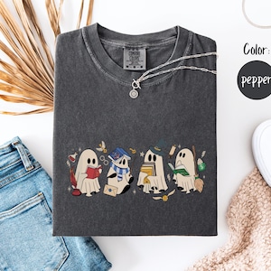May include: Pepper-colored t-shirt featuring a whimsical design of four cartoon ghosts, each with unique accessories like books, scarves, and hats. The shirt is styled with blue jeans, a silver necklace, and white sneakers. The color is pepper.