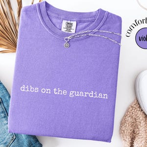May include: A violet short-sleeved t-shirt with a crew neckline featuring the white embroidered text "dibs on the guardian" across the chest. The shirt has a Comfort Colors brand label at the inner neck and a silver-toned chain necklace resting on top.