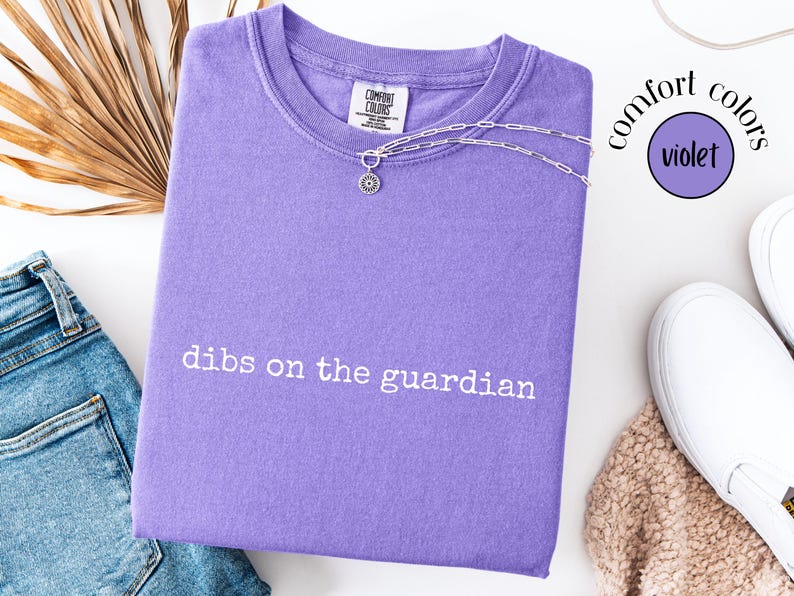 May include: A violet short-sleeved t-shirt with a crew neckline featuring the white embroidered text "dibs on the guardian" across the chest. The shirt has a Comfort Colors brand label at the inner neck and a silver-toned chain necklace resting on top.