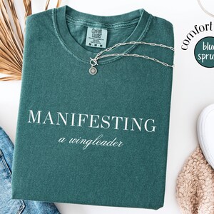 Op de afbeelding: Een blauwgroen Comfort Colors T-shirt met de woorden "MANIFESTING a wingleader" in het wit. Een zilveren ketting met een hanger ligt op het shirt. Het shirt ligt naast een spijkerbroek en witte sneakers.