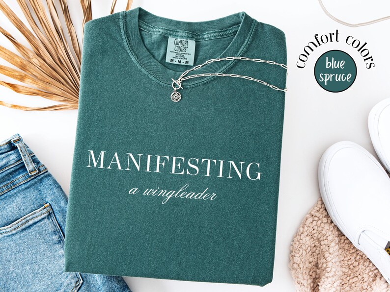 Op de afbeelding: Een blauwgroen Comfort Colors T-shirt met de woorden "MANIFESTING a wingleader" in het wit. Een zilveren ketting met een hanger ligt op het shirt. Het shirt ligt naast een spijkerbroek en witte sneakers.
