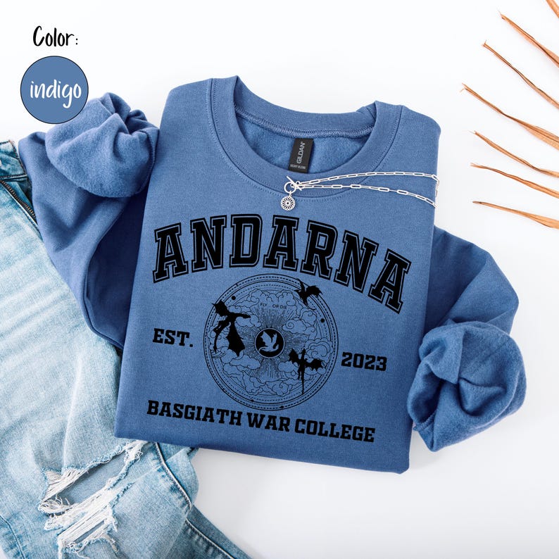 May include: Indigo blue sweatshirt with the text "ANDARNA" in black block letters. Below the text is a circular graphic with the words "EST. 2023" and "BASGIATH WAR COLLEGE". A silver necklace with a pendant is draped across the sweatshirt.