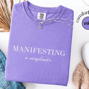 Op de afbeelding: Een violet Comfort Colors T-shirt met de woorden "MANIFESTING a wingleader" in het wit. Een zilveren ketting met een zonnehanger ligt op het shirt. Het shirt ligt naast een spijkerbroek, witte schoenen en een gedroogde plant.