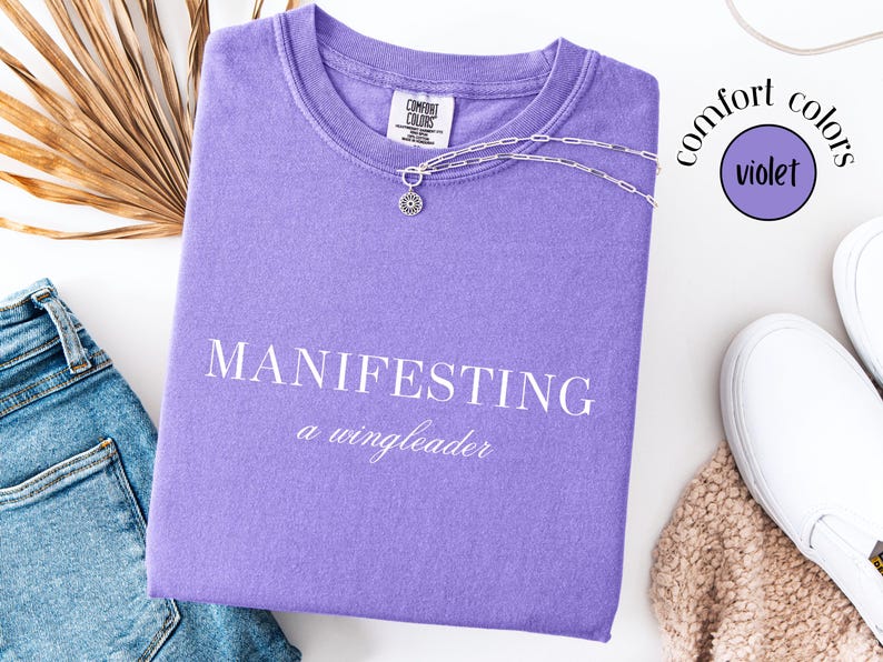 Op de afbeelding: Een violet Comfort Colors T-shirt met de woorden "MANIFESTING a wingleader" in het wit. Een zilveren ketting met een zonnehanger ligt op het shirt. Het shirt ligt naast een spijkerbroek, witte schoenen en een gedroogde plant.