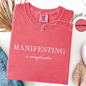 Op de afbeelding: Een koraalrood T-shirt met de witte tekst "MANIFESTING a wingleader". Een zilveren ketting met een hanger ligt op het shirt. Het shirt ligt naast een blauwe spijkerbroek en witte sneakers. Het shirt maakt deel uit van de "Comfort Colors" collectie.