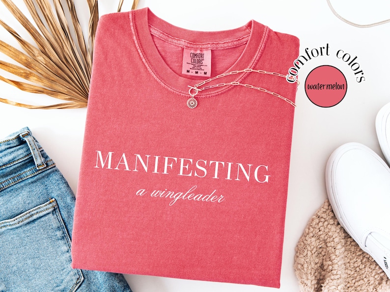 Op de afbeelding: Een koraalrood T-shirt met de witte tekst "MANIFESTING a wingleader". Een zilveren ketting met een hanger ligt op het shirt. Het shirt ligt naast een blauwe spijkerbroek en witte sneakers. Het shirt maakt deel uit van de "Comfort Colors" collectie.