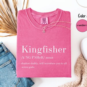 May include: A pink t-shirt with the word "Kingfisher" printed in white, along with a definition. A gold necklace with a pendant is on the shirt. The shirt is next to blue jeans, white shoes, and a dried palm leaf.