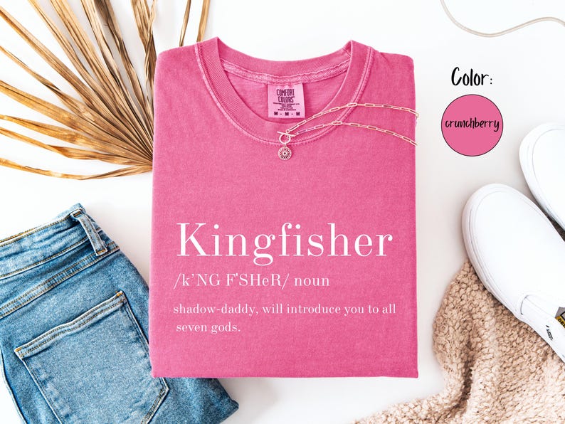 May include: A pink t-shirt with the word "Kingfisher" printed in white, along with a definition. A gold necklace with a pendant is on the shirt. The shirt is next to blue jeans, white shoes, and a dried palm leaf.