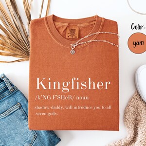May include: A yam-colored t-shirt with the word "Kingfisher" printed on the front. The shirt has a crew neck and short sleeves. A silver necklace with a pendant is draped on the shirt. Blue jeans and white shoes are also in the image.