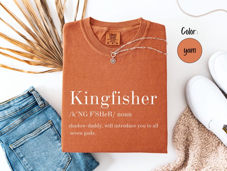 May include: A yam-colored t-shirt with the word "Kingfisher" printed on the front. The shirt has a crew neck and short sleeves. A silver necklace with a pendant is draped on the shirt. Blue jeans and white shoes are also in the image.