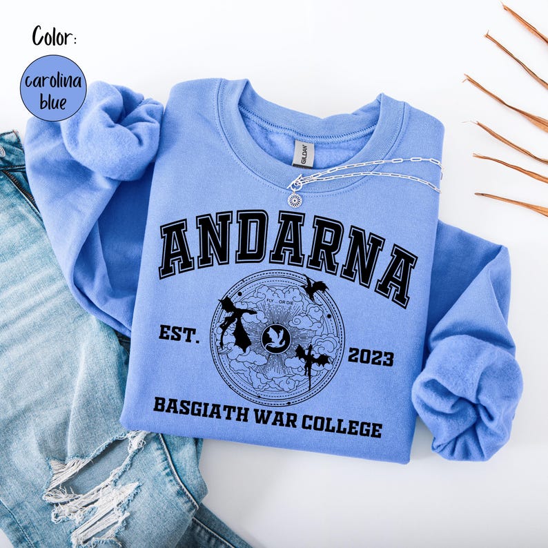 May include: Carolina blue sweatshirt with the word "ANDARNA" in black block letters. Below the word is a circular graphic with the text "EST. 2023 BASGIATH WAR COLLEGE". A silver necklace is draped across the sweatshirt.