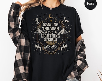 May include: Black long-sleeve shirt with a graphic design. The design features the text "DANCING THROUGH THE LIGHTNING STRIKES" with mountain and dragon illustrations. The shirt also has a crescent moon and stars.