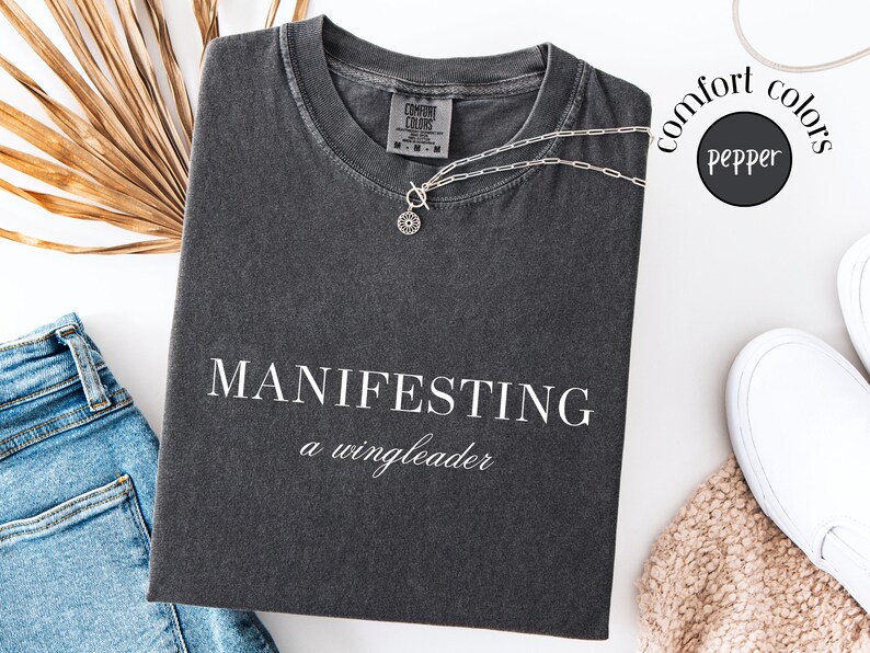 Op de afbeelding: Donkergrijs t-shirt met de witte woorden "MANIFESTING a wingleader". Een zilveren ketting met een cirkelvormige hanger ligt op het shirt. Het shirt ligt naast een spijkerbroek en witte sneakers.