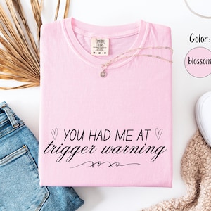 May include: A light pink t-shirt with the text "YOU HAD ME AT trigger warning xoxo" in black script. The shirt is folded and accessorized with a gold necklace and is next to blue jeans and white sneakers.