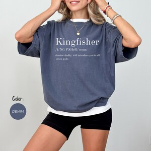 May include: Denim blue t-shirt with the word "Kingfisher" in white, along with a definition. The shirt has short sleeves and a relaxed fit. The text below the word "Kingfisher" reads "/K'NG F'SHER/ noun shadow-daddy, will introduce you to all seven gods."